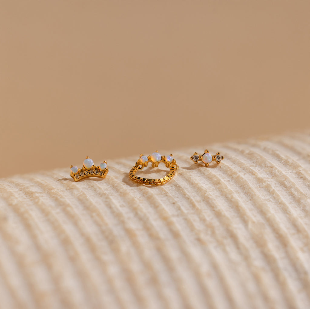 The Opal Cartilage Studs Set features three small gold crown-shaped rings, ideal for helix piercings, adorned with opals and gems, displayed on a textured beige surface.