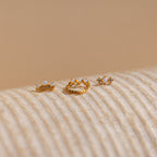 The Opal Cartilage Studs Set features three small gold crown-shaped rings, ideal for helix piercings, adorned with opals and gems, displayed on a textured beige surface.