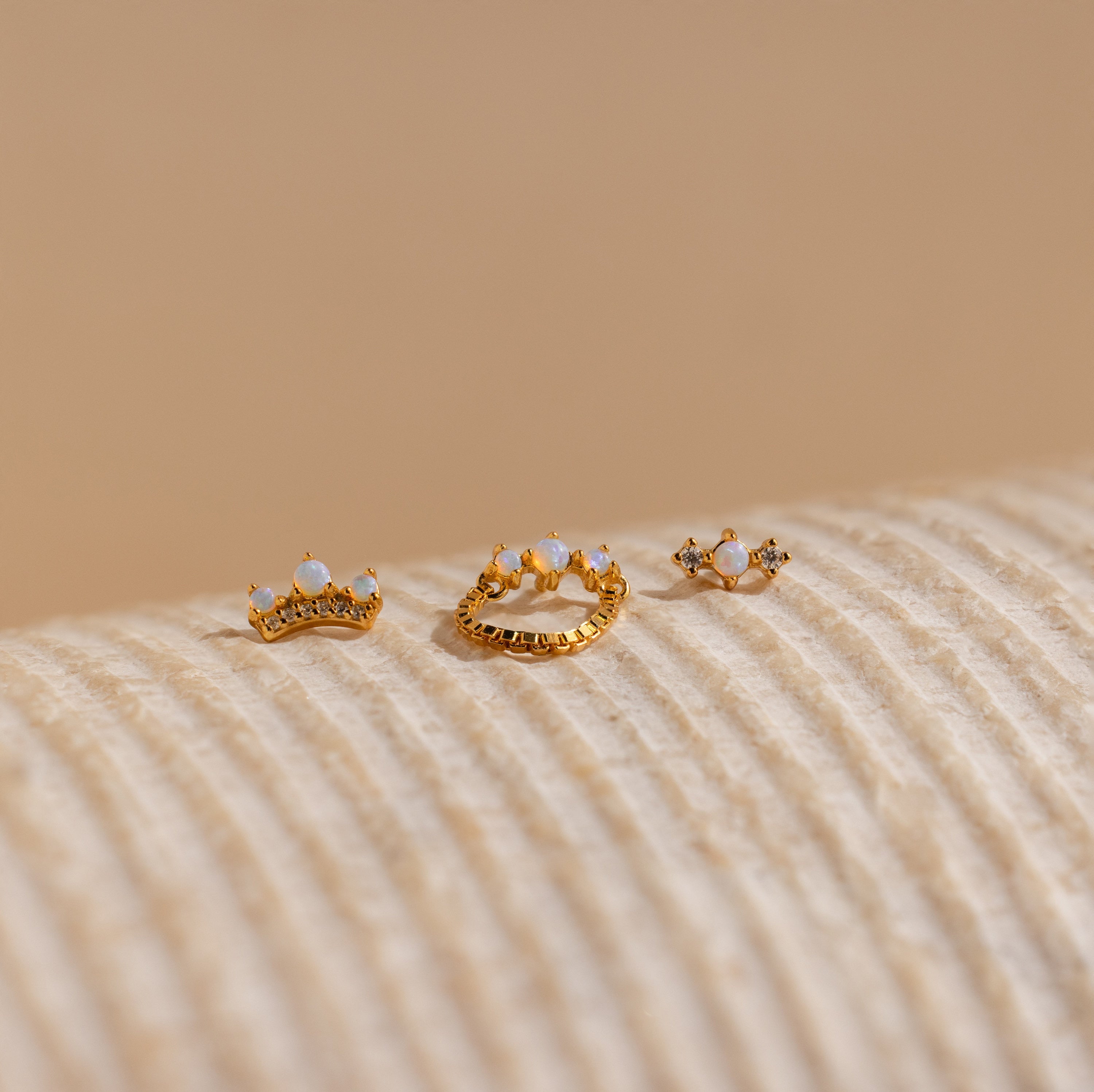 The Opal Cartilage Studs Set features three small gold crown-shaped rings, ideal for helix piercings, adorned with opals and gems, displayed on a textured beige surface.