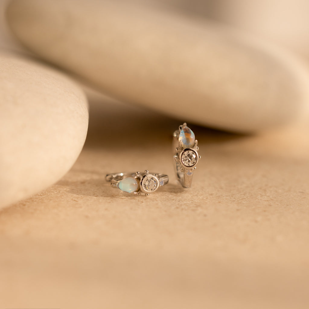 Two silver rings with gemstones rest on a smooth beige surface with rounded stones, perfectly paired with Opal and Diamond Huggies for a polished look.