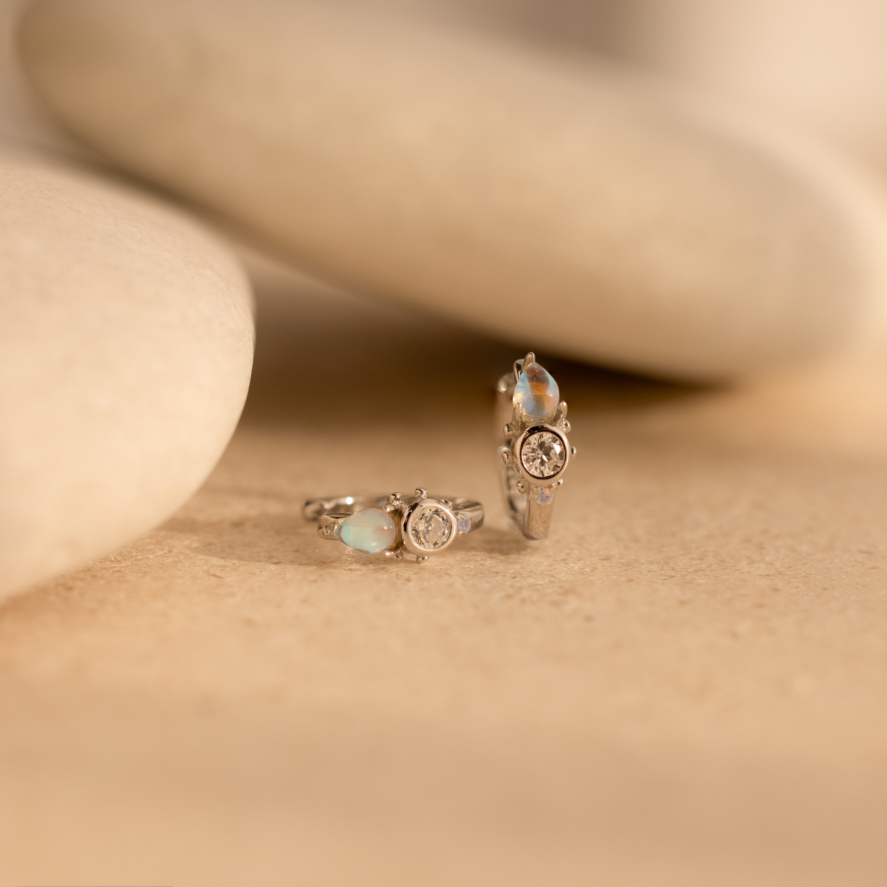 Two silver rings with gemstones rest on a smooth beige surface with rounded stones, perfectly paired with Opal and Diamond Huggies for a polished look.