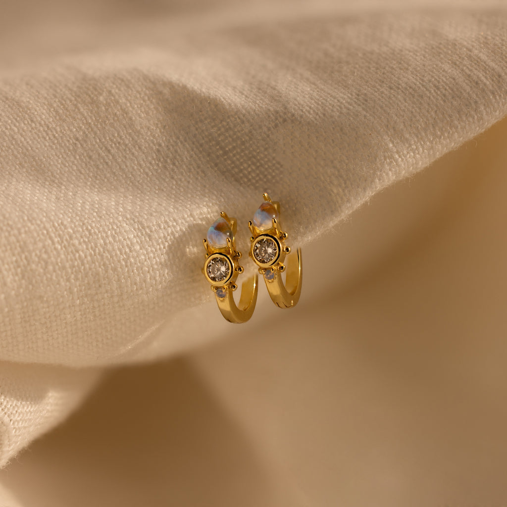 Opal and Diamond Huggies are elegant gold hoop earrings with gemstones, beautifully displayed on a soft beige fabric background.