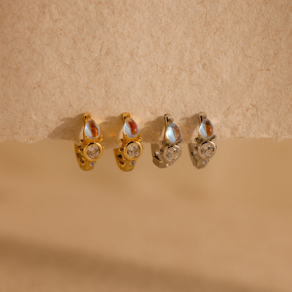Two pairs of gold and silver earrings—including Opal and Diamond Huggies and delicate hoops with round stones—rest on a textured beige surface.