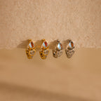 Two pairs of gold and silver earrings—including Opal and Diamond Huggies and delicate hoops with round stones—rest on a textured beige surface.