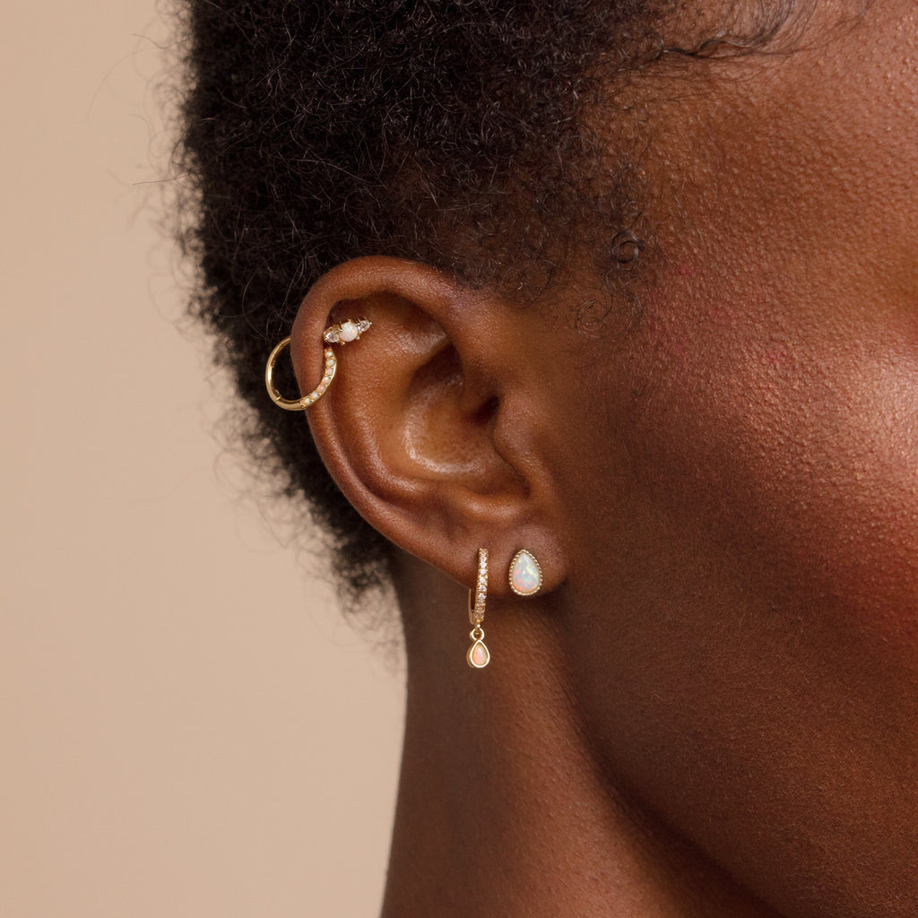 Close-up of a person's ear with three gold earrings, including a hoop and two dangle earrings—one pair is the Opal Teardrop Studs, bringing vintage-inspired elegance and gemstone sparkle for a standout jewelry look.