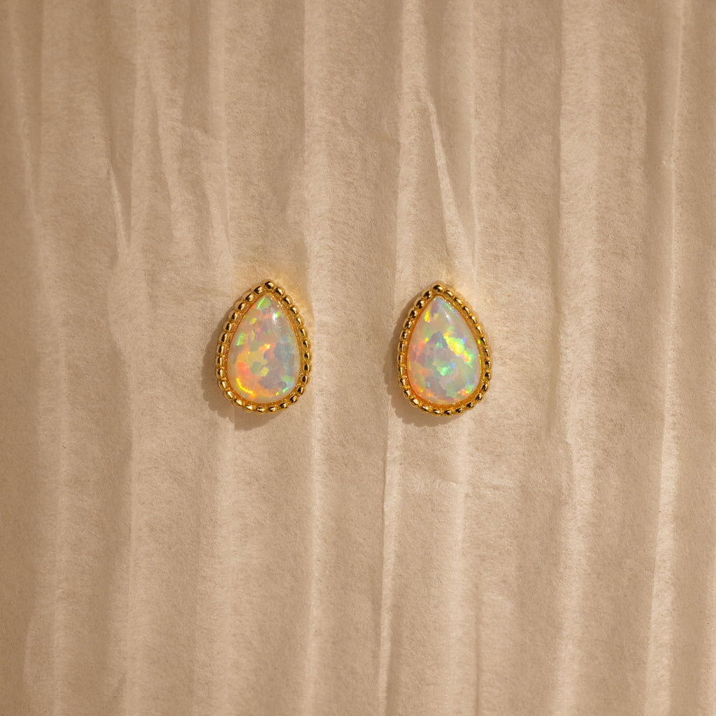 Opal Teardrop Studs shine on a textured beige backdrop—elegant, vintage-inspired jewelry with gold trim, perfect for making a statement.