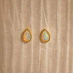 Opal Teardrop Studs shine on a textured beige backdrop—elegant, vintage-inspired jewelry with gold trim, perfect for making a statement.