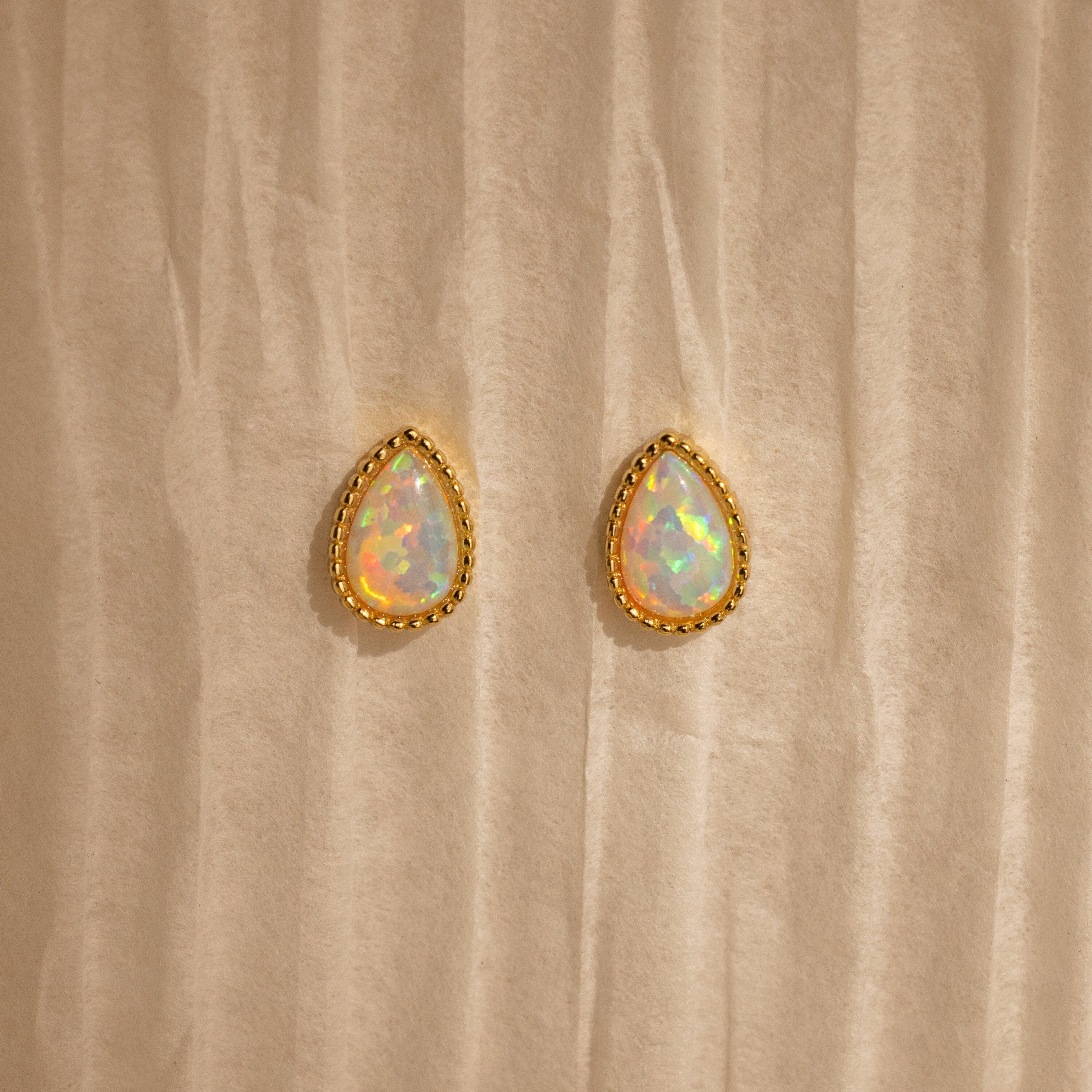 Opal Teardrop Studs shine on a textured beige backdrop—elegant, vintage-inspired jewelry with gold trim, perfect for making a statement.
