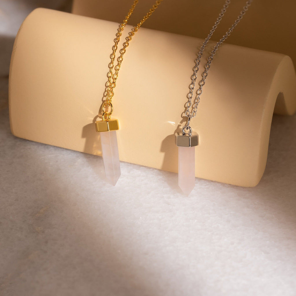 Crystal Quartz Necklaces—two hexagonal point pendants, one on a gold chain and one on silver, beautifully displayed on a beige stand—add positive energy and elegance to your style.