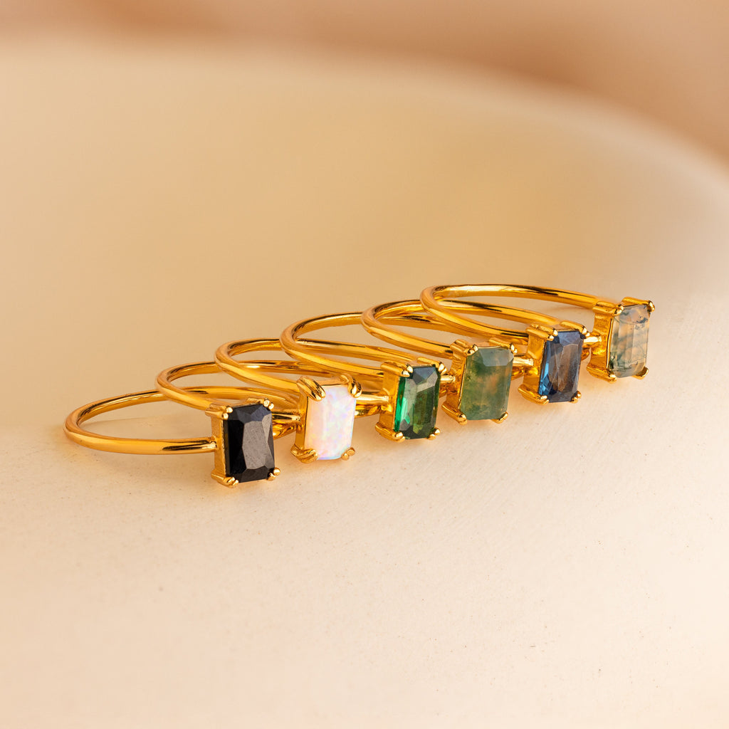 Six gold rings, featuring the Opal Emerald Ring, are displayed in a row on a soft beige surface—ideal as an engagement ring or a meaningful anniversary gift.