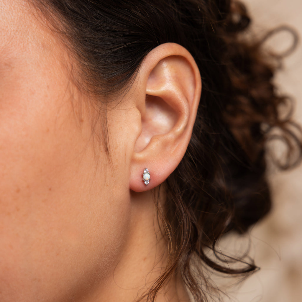 Close-up of a woman's ear wearing Opal and Diamond Studs, delicate three-stone earrings perfect for helix or cartilage piercings, featuring shimmering opal and diamond gemstones.