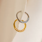 The Opal Cartilage Huggies—one silver with blue opals, one gold with white opals—rest on a soft beige surface. These minimalist earrings add subtle elegance to any look and are perfect for styling a helix piercing.