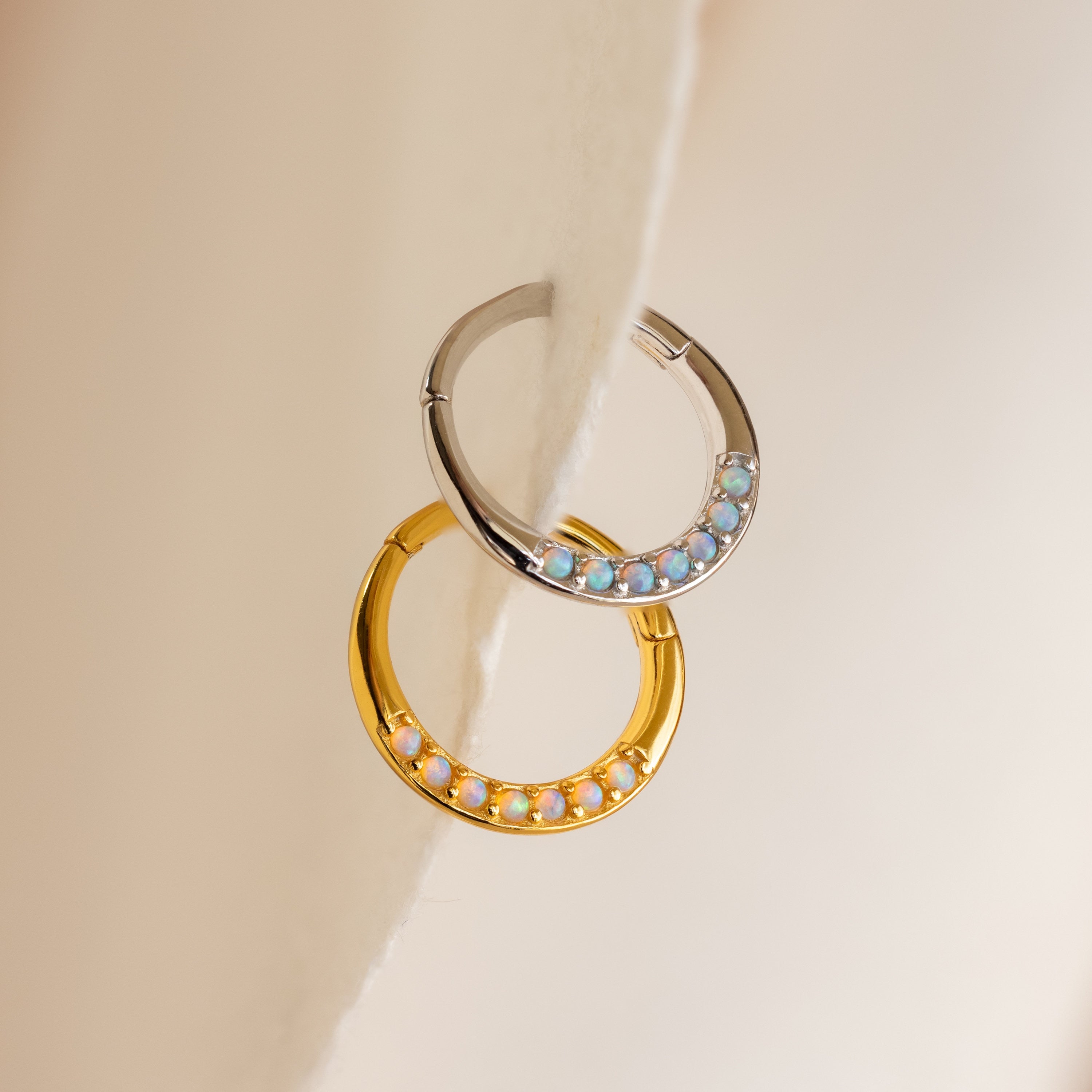 The Opal Cartilage Huggies—one silver with blue opals, one gold with white opals—rest on a soft beige surface. These minimalist earrings add subtle elegance to any look and are perfect for styling a helix piercing.
