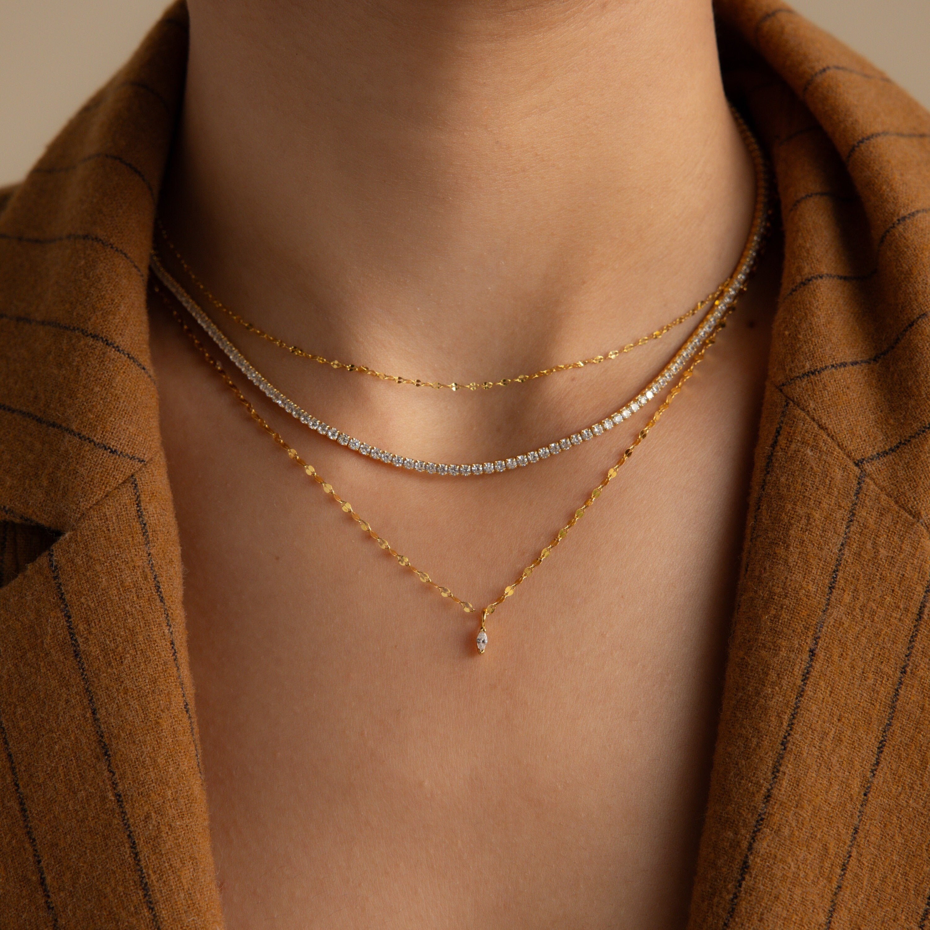 A person wearing three layered gold and silver necklaces, featuring the Dainty Marquise Pendant Necklace, paired with a brown pinstripe blazer.