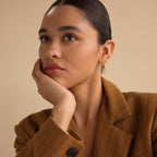 A woman in a brown pinstripe blazer, resting her chin on her hand and gazing thoughtfully to the side, wears Dainty Diamond Drop Earrings—an elegant choice for weddings or bridesmaid jewelry.