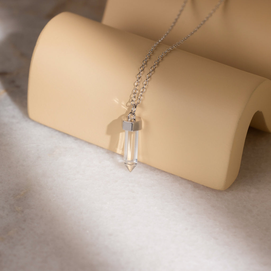 The Clear Quartz Necklace features a silver chain and a hexagonal clear quartz pendant, displayed on a curved beige stand—a timeless piece of crystal jewelry.