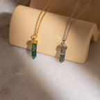 The gold Agate Quartz Necklace and a silver crystal pendant, both with green gemstone charms, are displayed on a curved beige stand in sunlight.