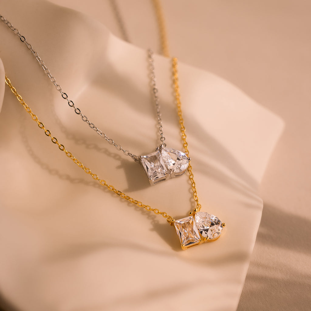 The Toi et Moi Diamond Necklace features two elegant pendants—one silver with an emerald-cut diamond, and one gold with a teardrop-shaped diamond—displayed on a soft background.