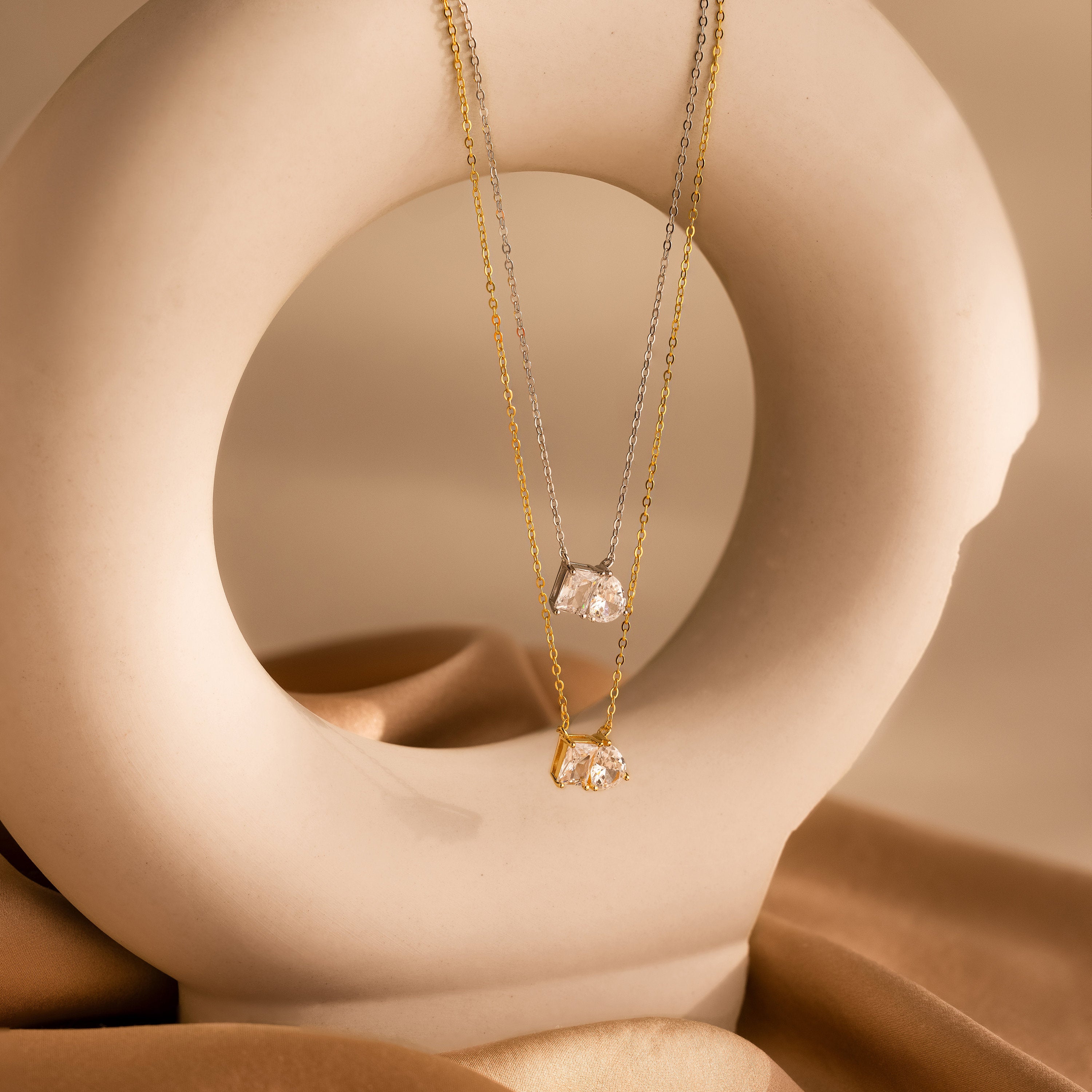 The Toi et Moi Diamond Necklace comes in both gold and silver, featuring a sparkling teardrop-shaped diamond, elegantly displayed on a smooth, circular beige sculpture.