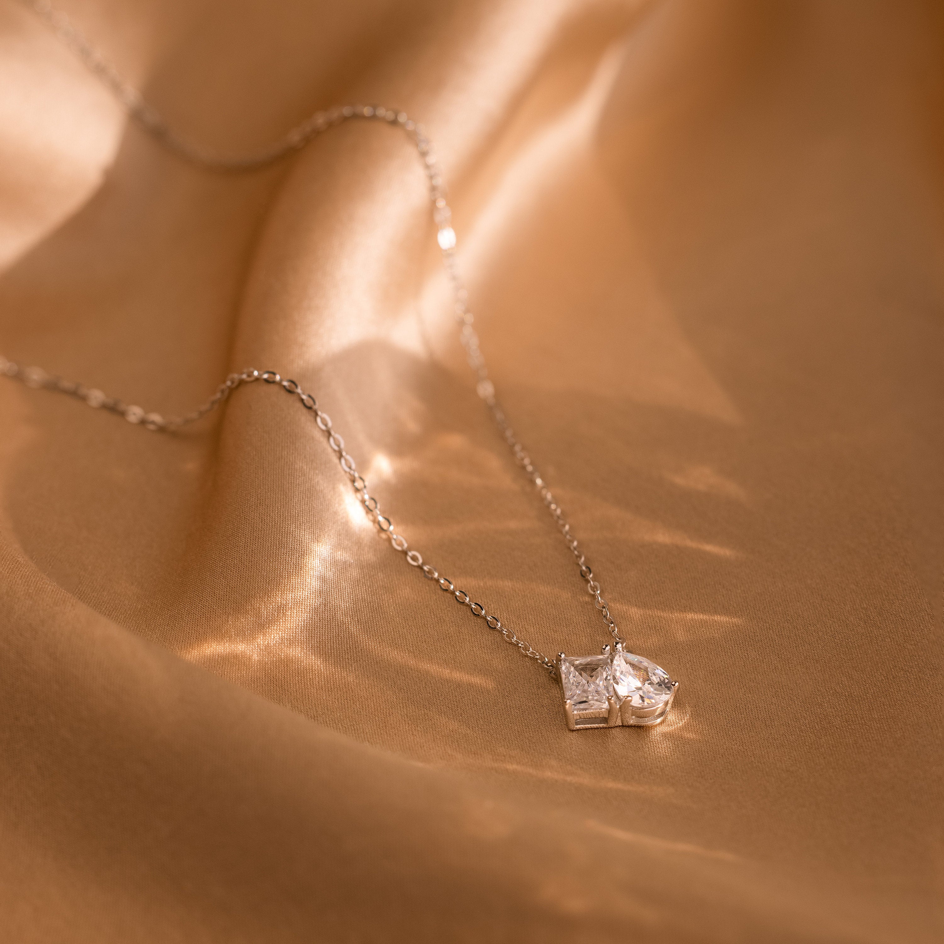 The Toi et Moi Diamond Necklace, featuring two square gemstones, is displayed on smooth, golden fabric.