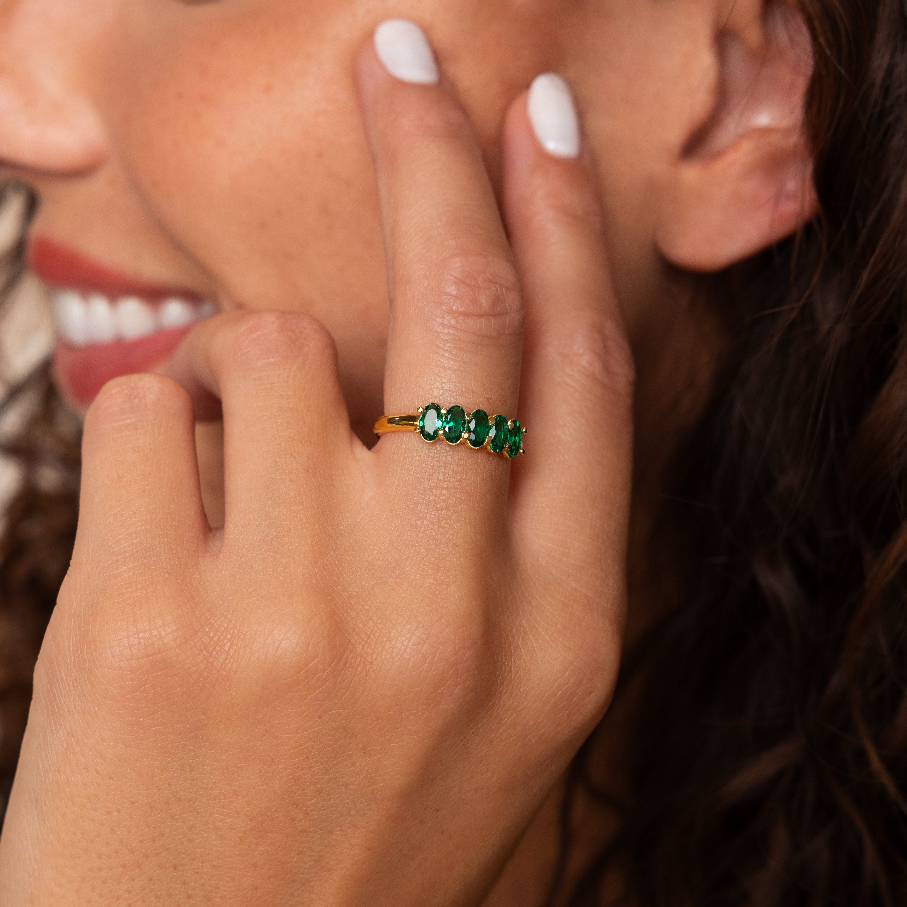 A woman with curly hair smiles while wearing the Oval Birthstone Ring featuring elegant green gemstones on her finger.