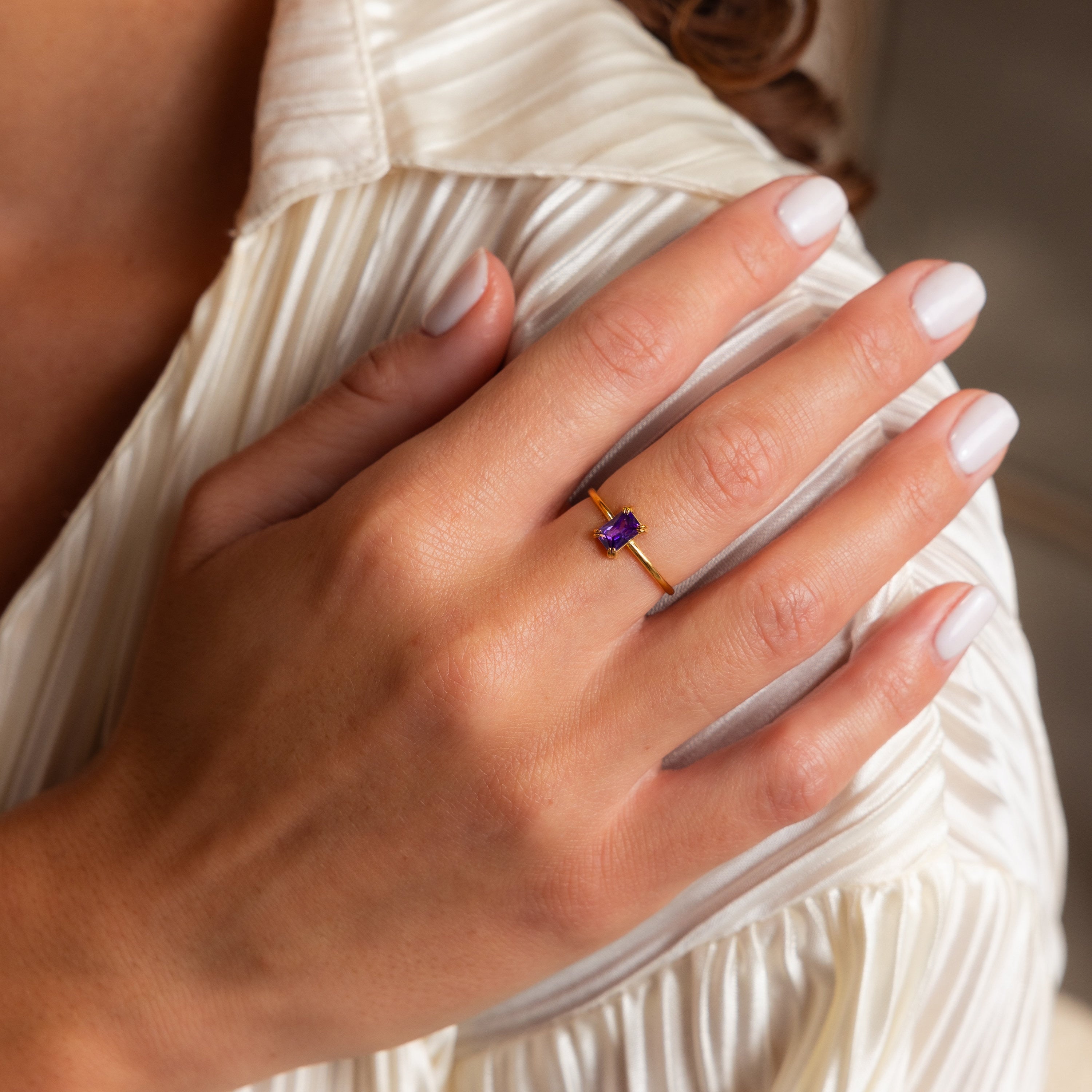A hand with white nails wears the Emerald Cut Birthstone Ring, showcasing a rectangular purple gemstone atop a pleated blouse—an elegant choice for a Christmas gift.