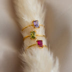 Three Emerald Cut Birthstone Rings in gold, each with a square purple, green, or red gemstone, displayed on soft cream fabric—perfect for Christmas or as a meaningful birthstone gift.