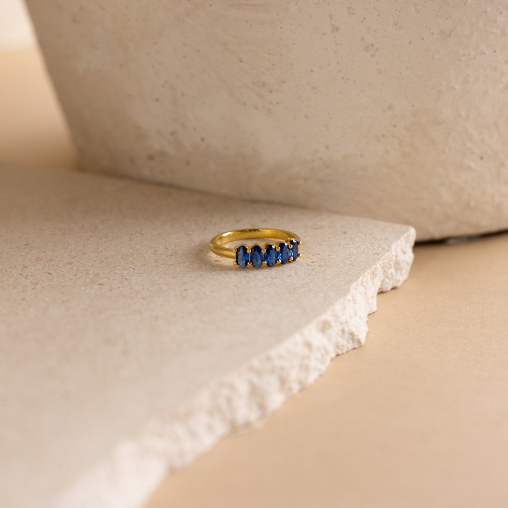 The Oval Birthstone Ring, adorned with five blue gemstones, is displayed on a textured, off-white stone surface.