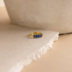 The Oval Birthstone Ring, adorned with five blue gemstones, is displayed on a textured, off-white stone surface.