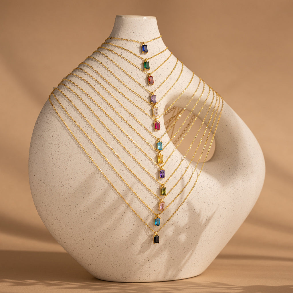 The Emerald Cut Birthstone Necklace features a vibrant gemstone pendant on a gold chain, elegantly displayed over a sculptural white vase with a beige backdrop—a customizable necklace that makes the perfect Christmas gift.