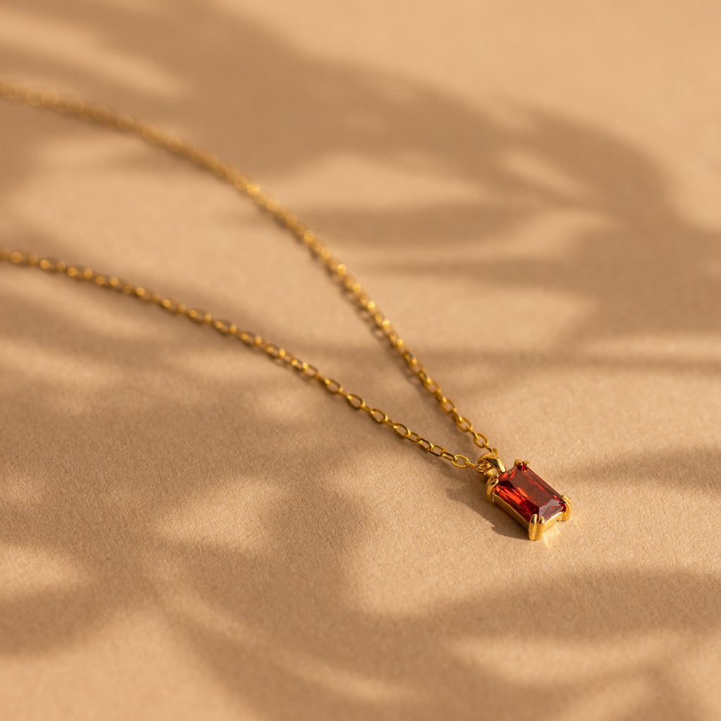 The Emerald Cut Birthstone Necklace features a gold chain with a red emerald-cut gemstone pendant, showcased on a beige background with soft shadows—an ideal Christmas gift.