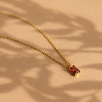 The Emerald Cut Birthstone Necklace features a gold chain with a red emerald-cut gemstone pendant, showcased on a beige background with soft shadows—an ideal Christmas gift.