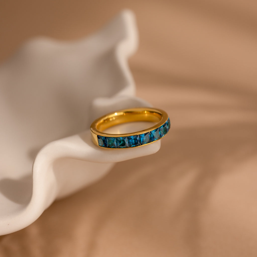 The Birthstone Inlay Band, featuring blue gemstones set in gold, rests on a wavy white ceramic dish against a soft beige background, making it a striking statement accessory.