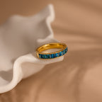 The Birthstone Inlay Band, featuring blue gemstones set in gold, rests on a wavy white ceramic dish against a soft beige background, making it a striking statement accessory.