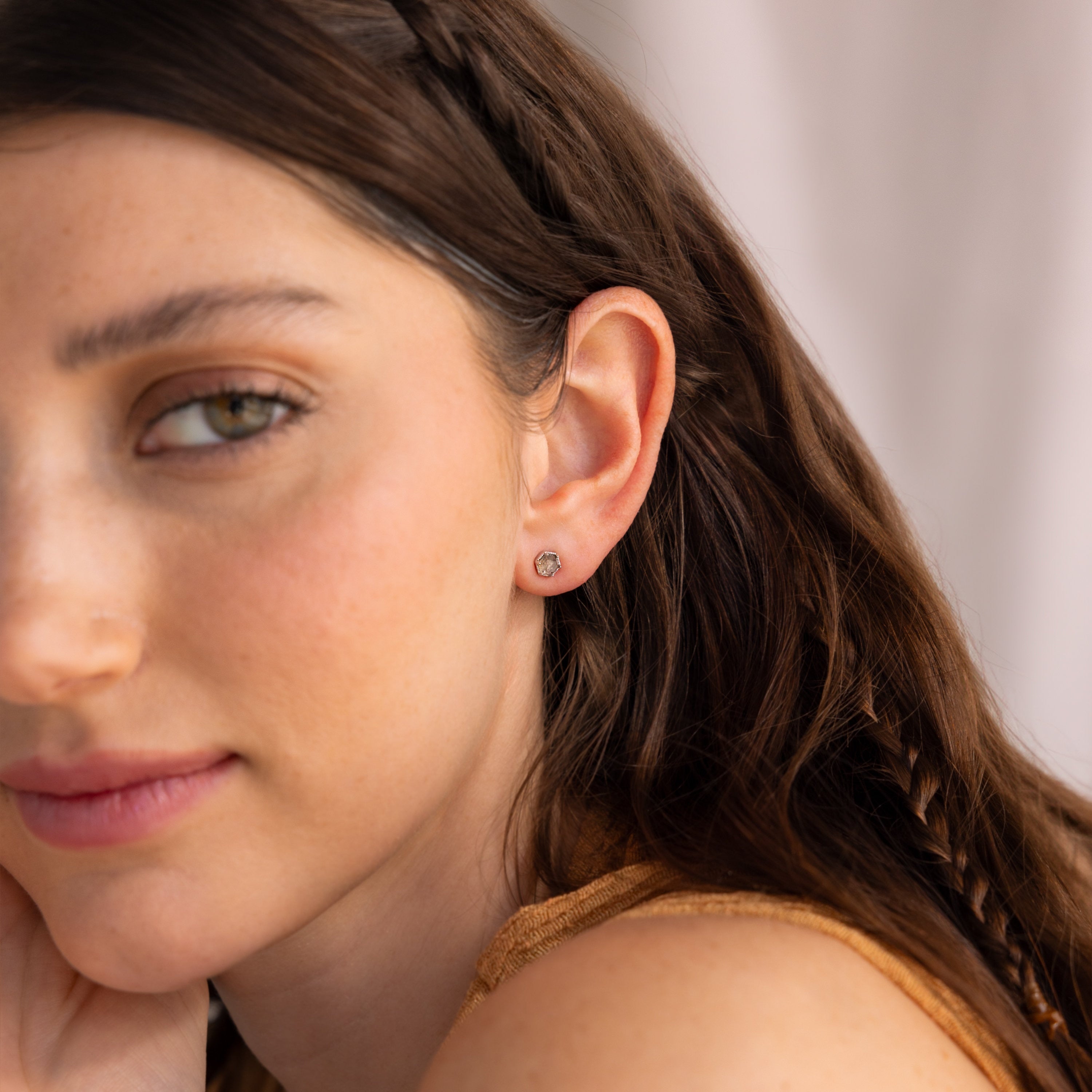 A woman with long brown hair, wearing Labradorite Hexagon Studs, looks slightly to the side.