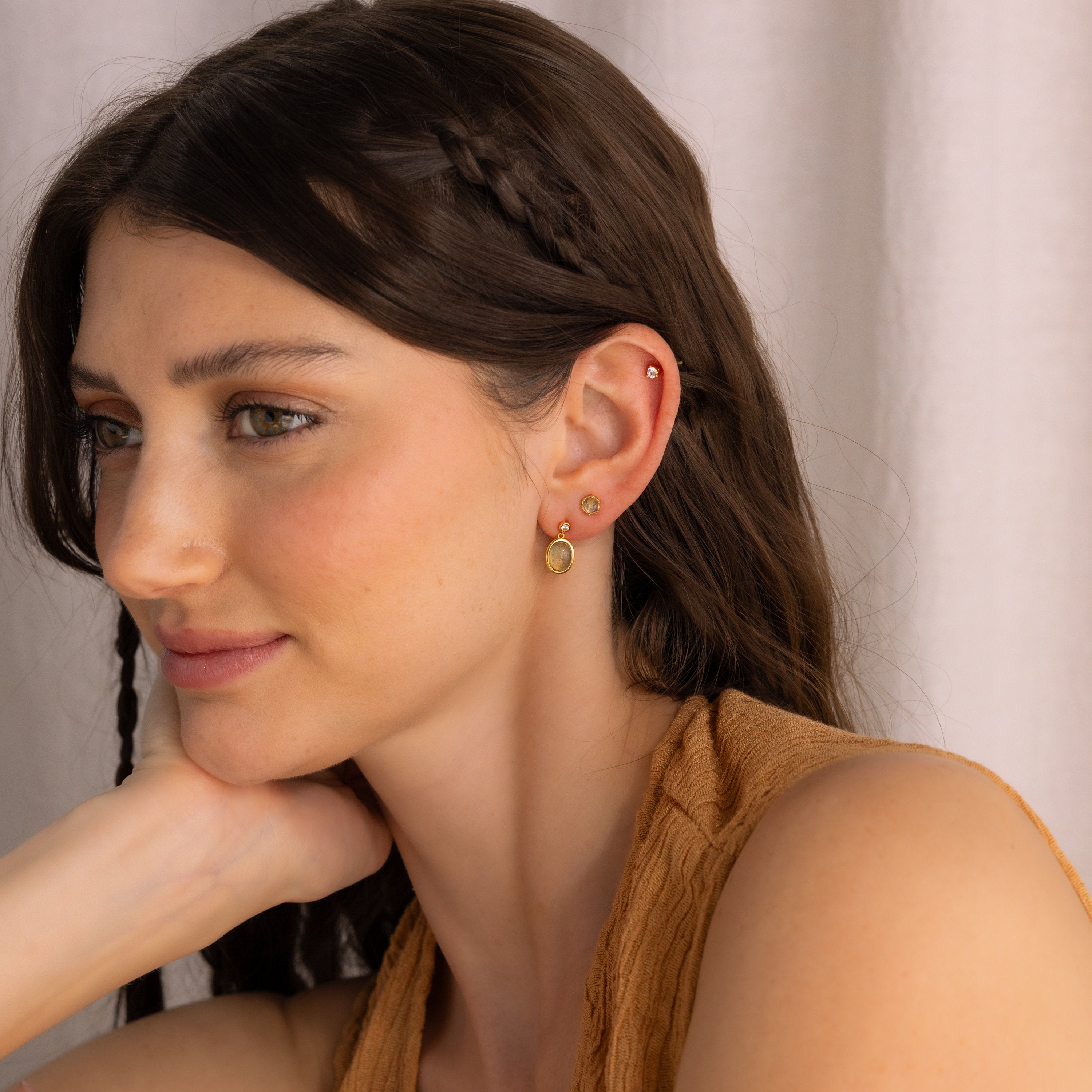 A woman with long brown hair and a braid smiles softly, resting her chin on her hand while wearing Labradorite Hexagon Studs.