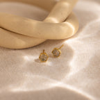 Labradorite Hexagon Studs with textured gold centers rest on a sandy surface beside a curved beige object—ideal for lovers of mystical jewelry or anyone seeking unique labradorite earrings.