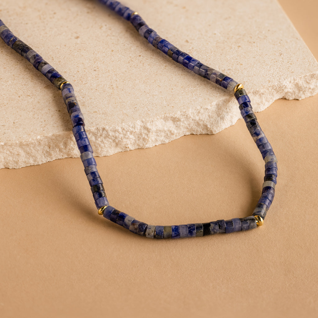 The Tanzanite Beaded Necklace, accented with gold spacers and vibrant blue beads, is beautifully showcased on a beige stone and tan background—ideal for fans of mystical jewelry.