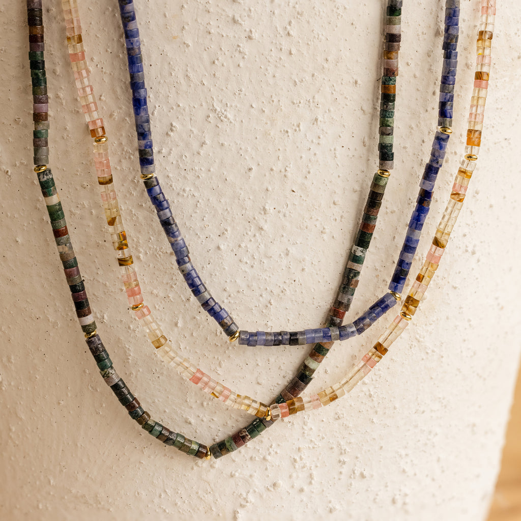 The Tanzanite Beaded Necklace features shimmering tanzanite blue beads and gold accents, creating a mystical jewelry charm when displayed against a textured white surface.