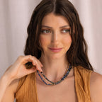 A woman with long brown hair smiles softly, wearing a sleeveless brown top and layered mystical jewelry, featuring the Tanzanite Beaded Necklace.