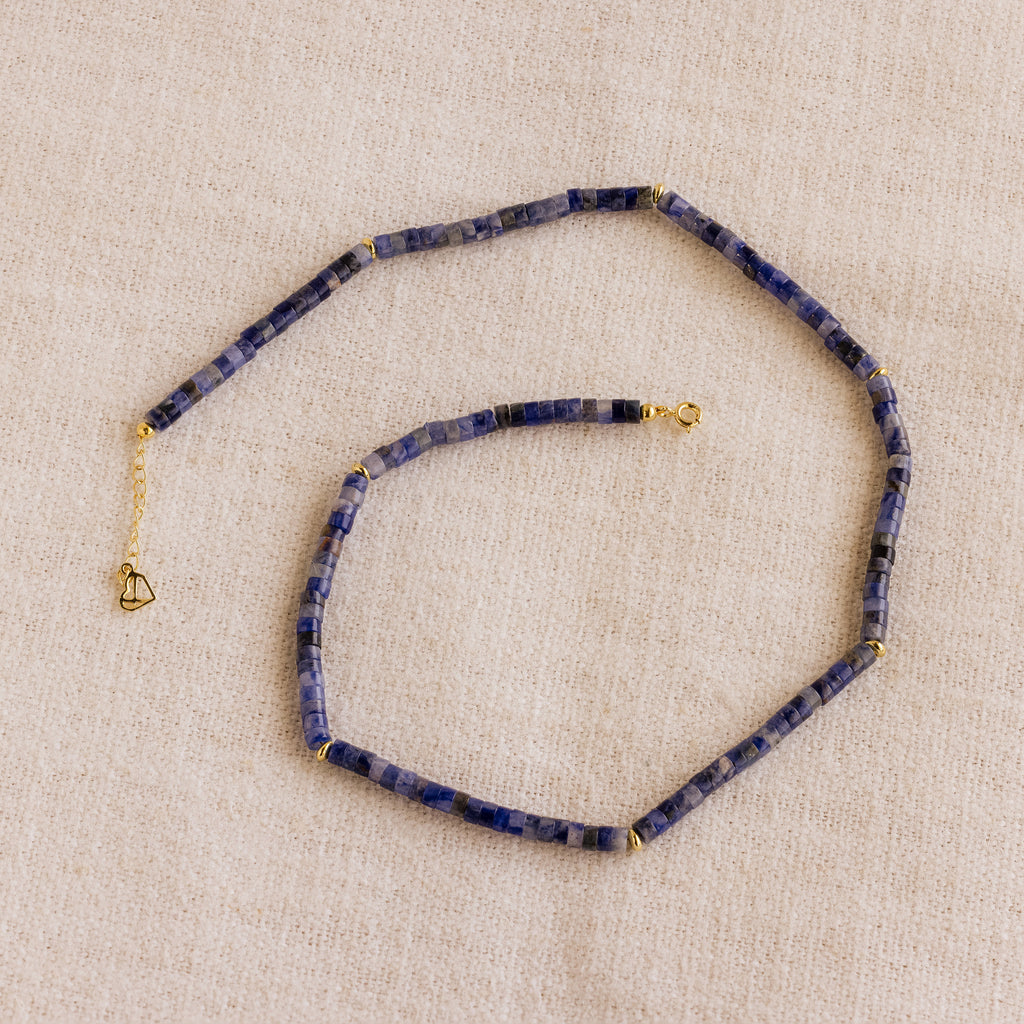 The Tanzanite Beaded Necklace, featuring blue stones and gold accents, is elegantly showcased on beige fabric—ideal for those seeking mystical jewelry.