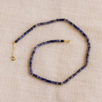 The Tanzanite Beaded Necklace, featuring blue stones and gold accents, is elegantly showcased on beige fabric—ideal for those seeking mystical jewelry.