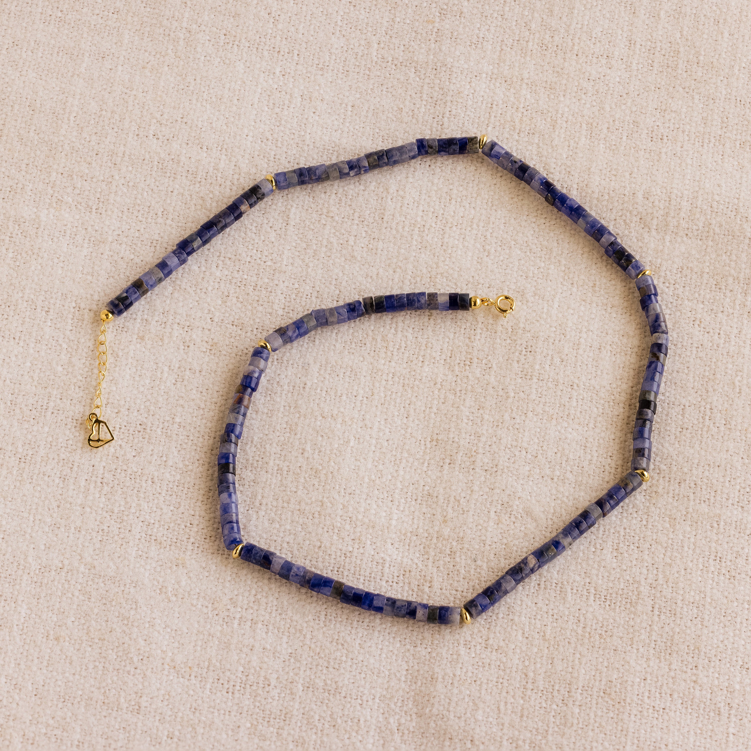 The Tanzanite Beaded Necklace, featuring blue stones and gold accents, is elegantly showcased on beige fabric—ideal for those seeking mystical jewelry.