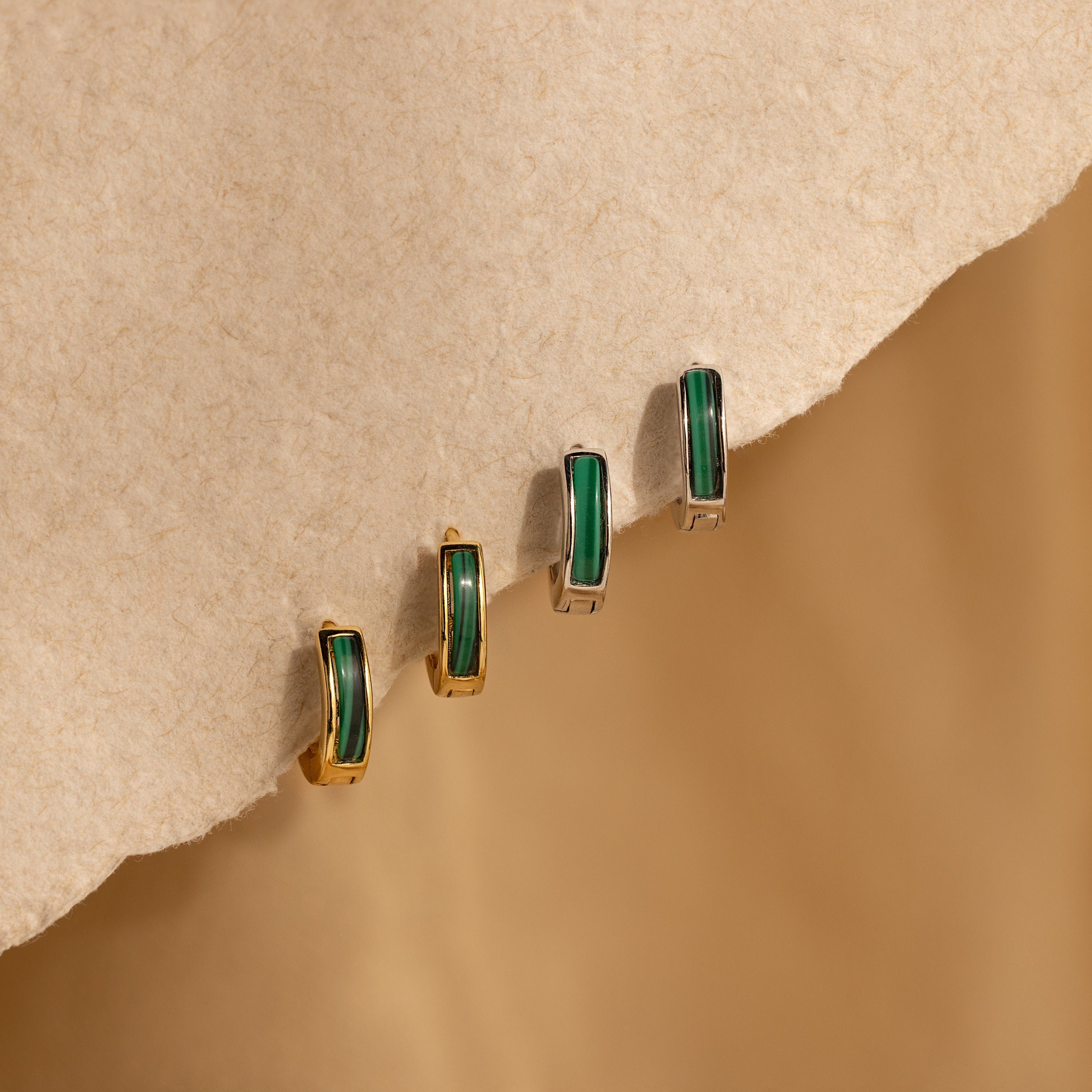 Two pairs of Malachite Inlay Huggies earrings in gold and silver are displayed on a textured beige surface.