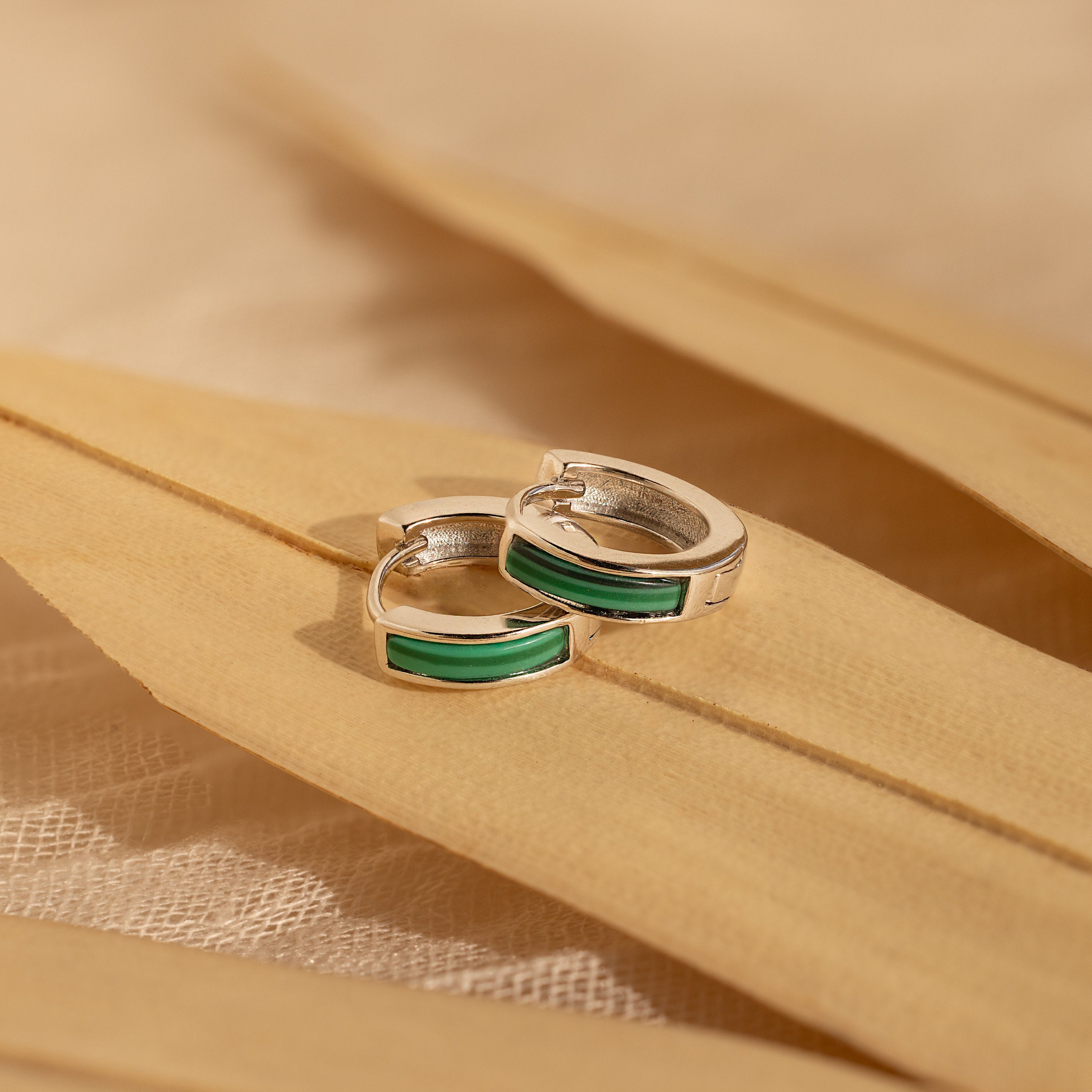 Malachite Inlay Huggies featuring silver hoops and green malachite inlay are displayed on beige dried leaves, captured in soft natural light.
