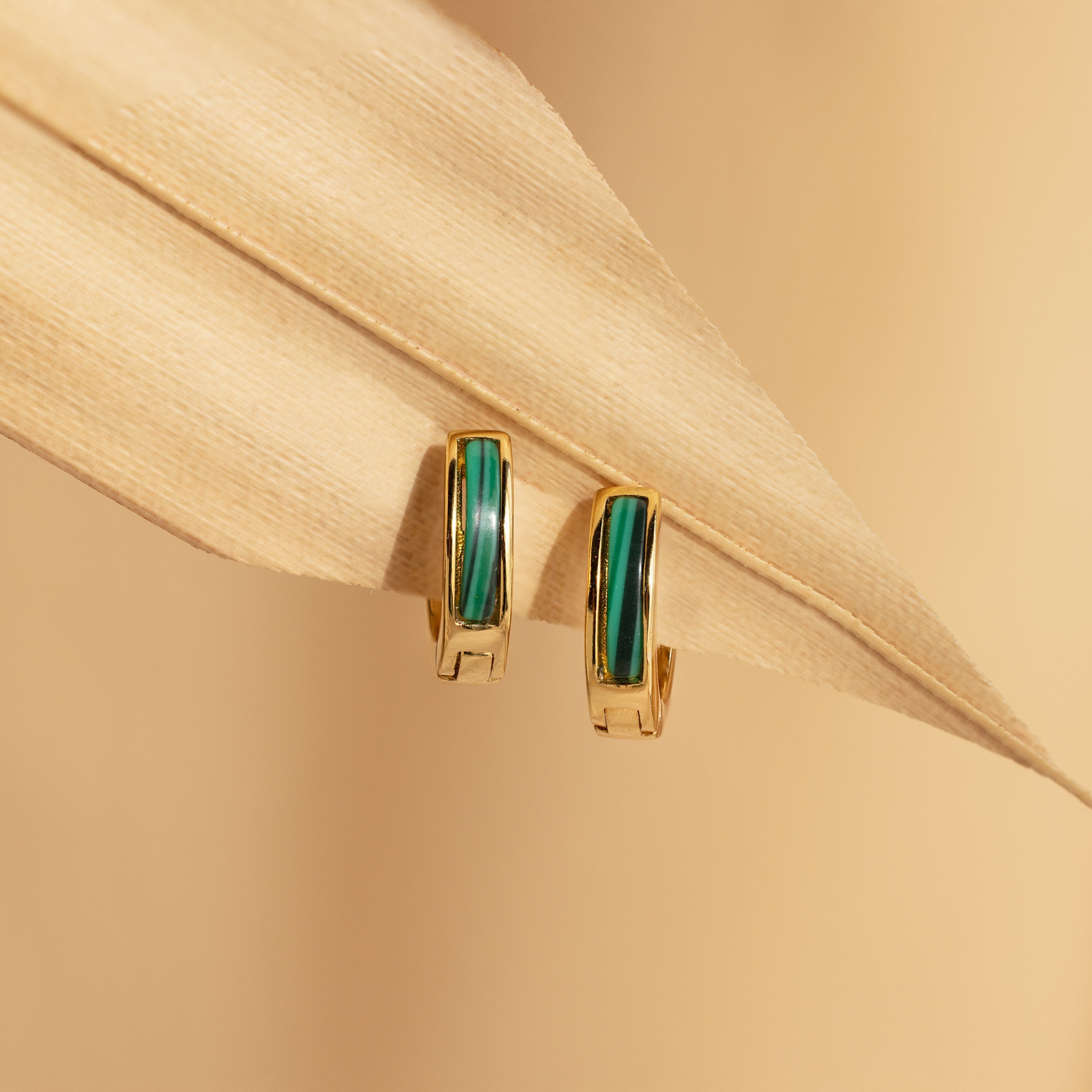 Malachite Inlay Huggies featuring vibrant green malachite rest on a beige dried leaf against a soft tan background, emphasizing the elegance of these distinctive green stone earrings.