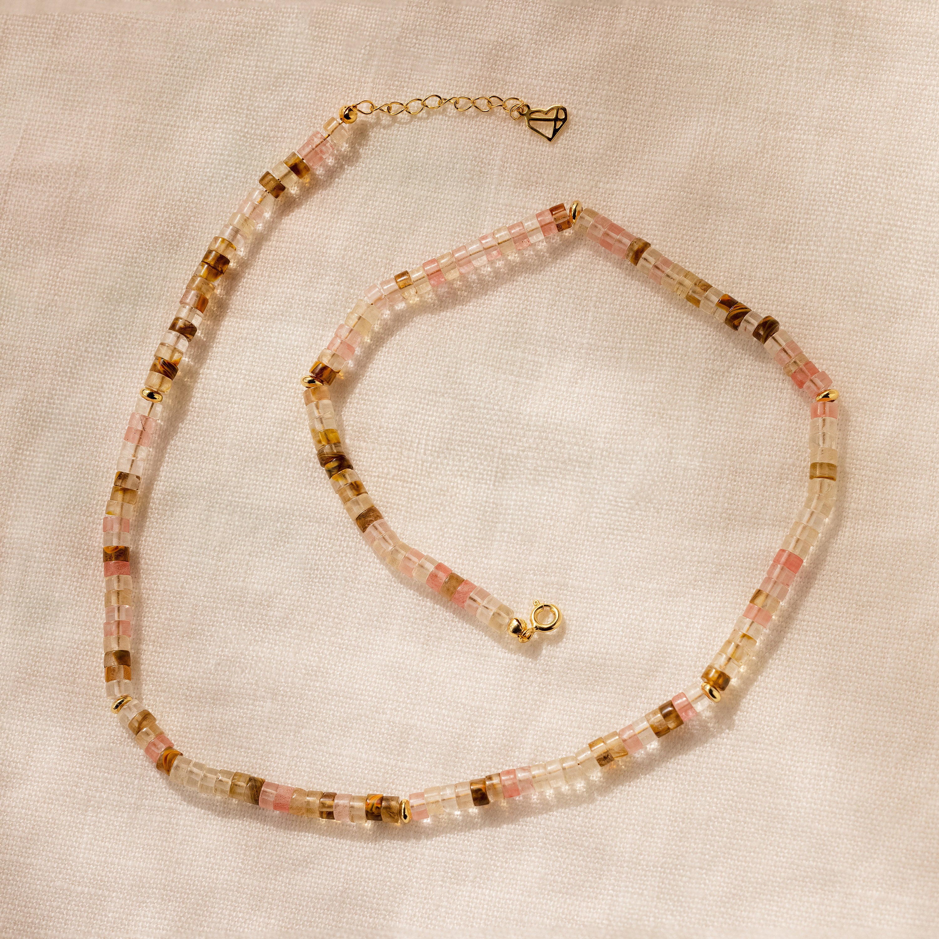 The Rose Quartz Beaded Necklace, with pink, clear, and gold square rose quartz beads, is displayed on a light fabric—an elegant gift for your best friend.