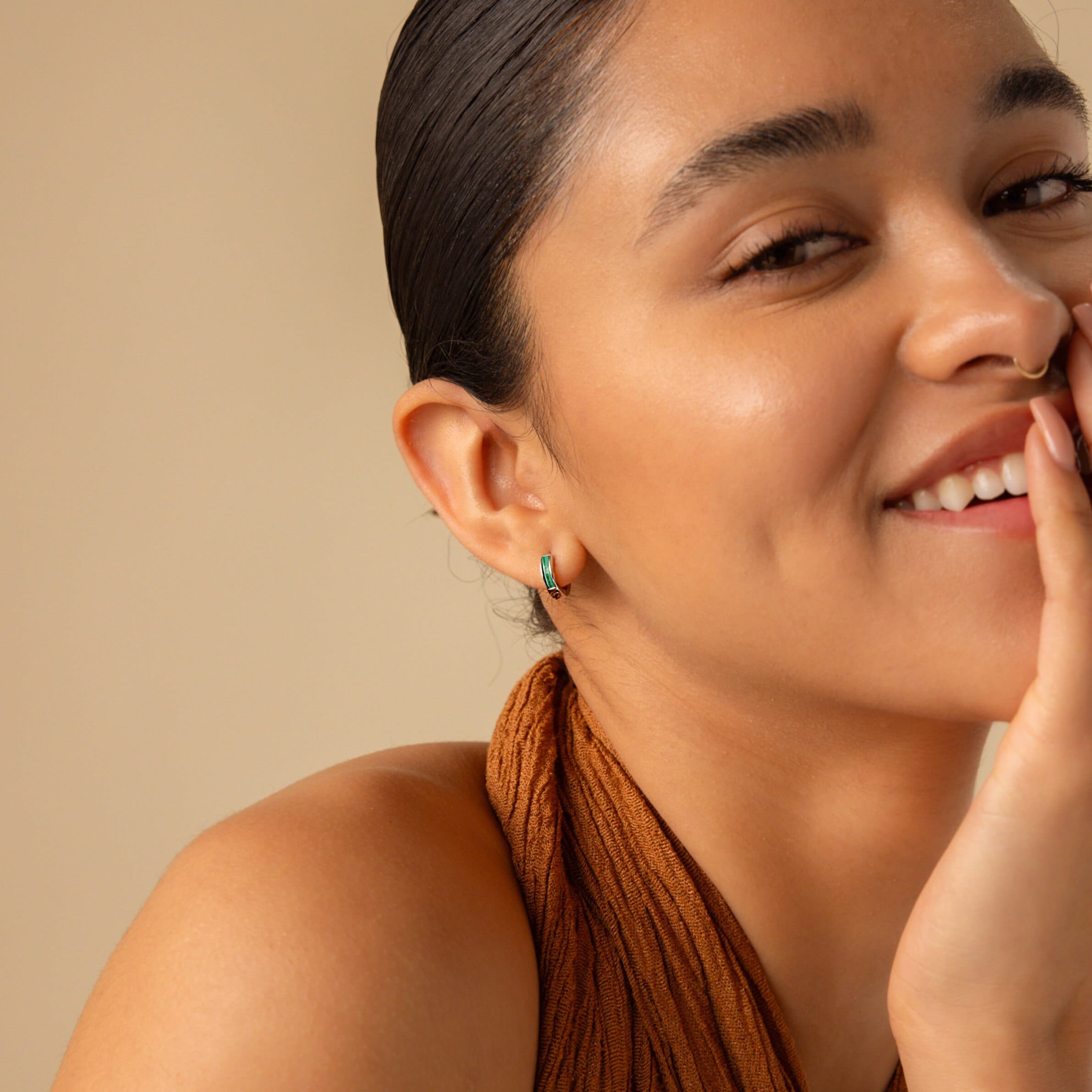 A smiling woman with slicked-back hair wears a brown top and Malachite Inlay Huggies, set against a beige background.