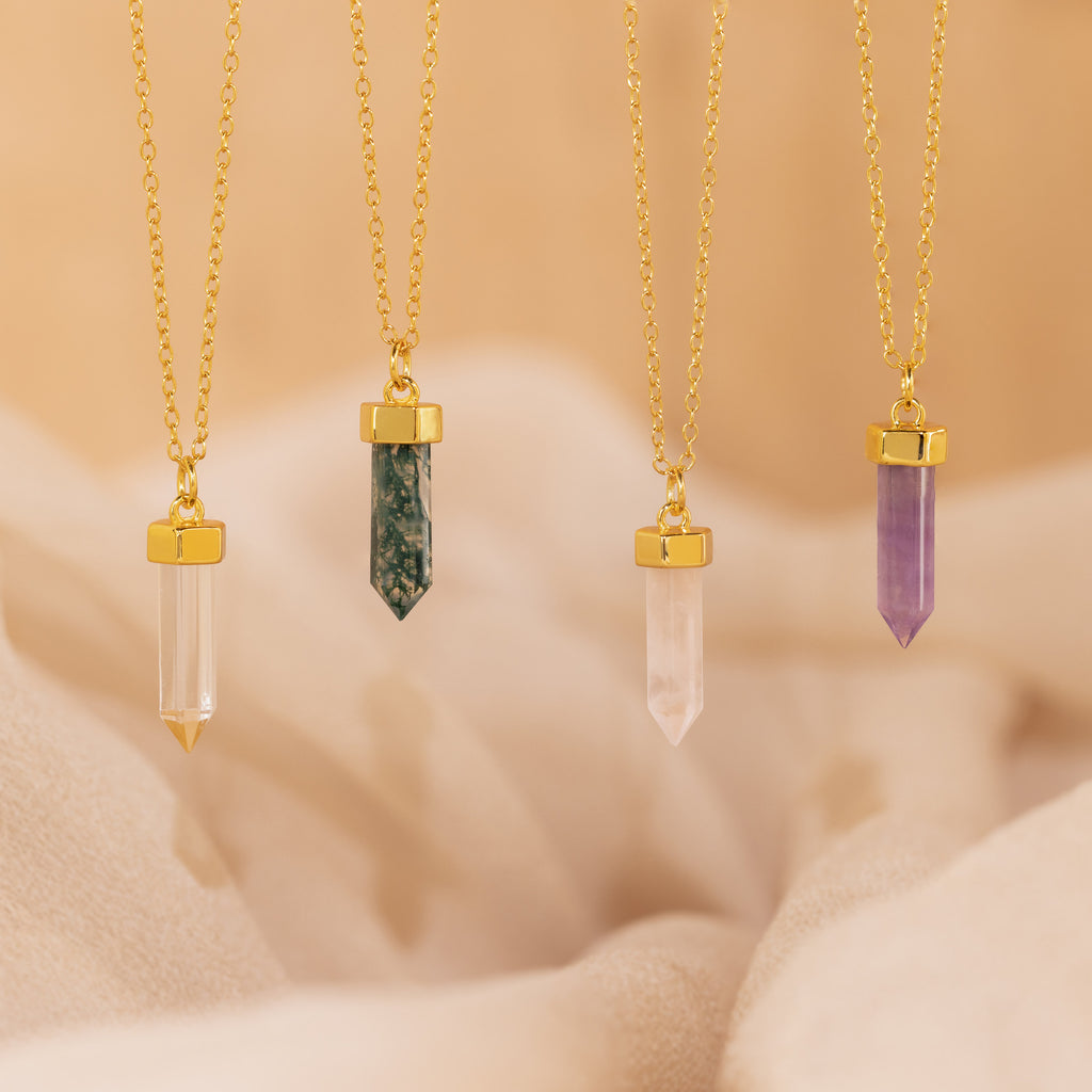 Four Crystal Quartz Necklaces, each featuring a gold chain and a hexagonal point pendant in clear, green, pink, or purple, rest on a soft beige background—designed to radiate positive energy.