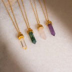 Four gold-chained Crystal Quartz Necklaces in hexagonal point designs—yellow, green, pink, and purple—rest on a light textured surface, radiating positive energy.